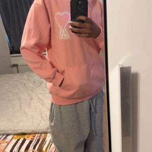 Rosa hoodie från Ami med hjärtdetalj - Säljer en snygg rosa hoodie från Ami med broderad hjärtdetalj och bokstaven A på bröstet. Hoodien har huva med snörning, stor magficka och långa ärmar. Perfekt för dig som gillar chill och stilren streetwear.