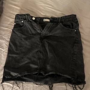 Svart jeanskjol från Gina  - Svart jeanskjol med rå kant och slitna detaljer från g perfect jeans. Klassisk femficksmodell med dragkedja och knapp framtill. Kjolen är kort och har en cool, avslappnad vibe som passar perfekt till sneakers eller boots.