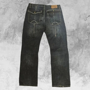 Rare T&K Dark Wash Bootcut Jeans - T&K JEANS i en snygg mörk tvätt med coola detaljer. Lätt slitning på låren och en klassisk bootcut-passform som sitter perfekt över skorna. Bakfickor med unik lockdesign och vertikal söm, plus ett läderpatch i midjan. Litet hål på vänster sida, knappt synligt vid användning, och det är även inte möjligt att helt stänga en av bakfickorna som det visas i bilden..  Mått: Längd – 111 cm Midja – 44,5 cm Benslut – 23 cm  Ett kvalitetsfynd, passa på innan de försvinner!