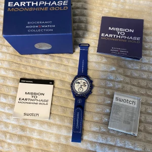 Omega x Swatch MoonSwatch Earthphase - Säljer en Omega x Swatch MoonSwatch Earthphase Mission to Earthphase Moonshine Gold. Klockan har en blå biokeramisk boett, vit urtavla med guld- och blå detaljer, samt blå textilrem med Speedmaster- och MoonSwatch-logga. Baksidan har en cool jordglobsgrafik.
