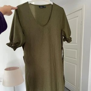 Olivgrön kort klänning från Esmara - Olivgrön klänning från Esmara med v-ringning och kort puffärm med resår. Materialet är lätt strukturerat och har en mjuk känsla. Modellen är rak och kort, med knytband i sidan för extra detalj. Perfekt för dig som gillar enkel men snygg stil.