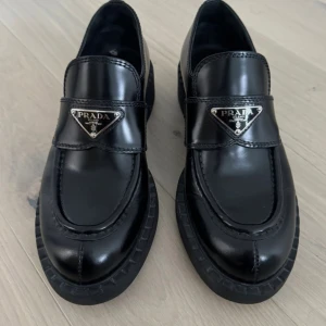 Svarta loafers från Prada i skinn - Snygga svarta loafers från Prada i glansigt skinn med chunky sula och ikonisk metallogo framtill. Skorna har rund tå och klassisk siluett som ger en stilren och lyxig vibe. Skorna e oanvända kommer med box säljer skorna åt min syster hon köpte dom på Plick för ett år sen men dom var för små och nu vill hon sälja dom hon vet ej om dom e äkta. Skriv vid intresse🤩