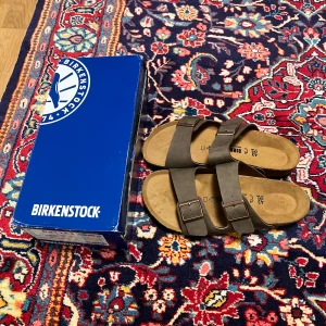 Birkenstock Arizona sandaler brun mocka - Säljer ett par Birkenstock Arizona sandaler i brun mocka med två breda remmar och justerbara metallspännen . Fotbädden är i kork och innersulan är mjuk och bekväm. Perfekta för sommaren och riktigt sköna att glida runt i. Lite stora passar bättre till en 44/45