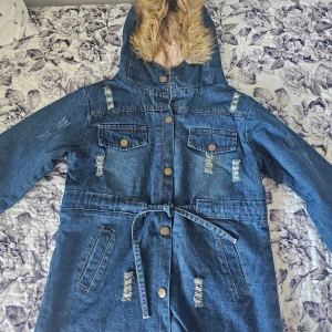 Blå jeansjacka med fuskpäls och bälte - Snygg blå jeansjacka med huva fodrad i beige fuskpäls. Jackan har slitna detaljer, knappar i metall, två bröstfickor och ett bälte i midjan. Långärmad och med coola slitningar som ger en trendig vibe. Perfekt för dig som gillar streetstyle.