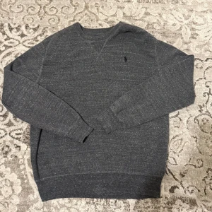 Mörkgrå stickad tröja Polo Ralph Lauren - Mörkgrå stickad tröja från Polo Ralph Lauren med diskret logga broderad på bröstet. Tröjan har rund hals, ribbade muddar och långärmad passform. Perfekt för dig som gillar klassisk och enkel stil med premiumkänsla.