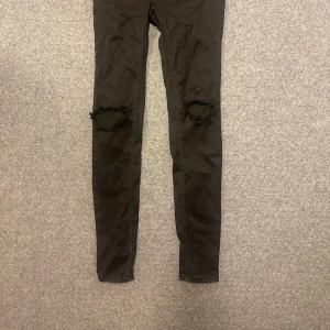 Svarta skinny jeans med hål på knäna - Svarta skinny jeans med hög midja och slitna hål på båda knäna. Jeansen har klassisk femficksdesign och smal passform hela vägen ner. Perfekta för en edgy och trendig look.