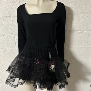 Svart tyllkjol med stjärnor Living Dead Souls - Cool svart kortkjol från Living Dead Souls med flera lager tyll och spets. Kjolen har färgglada stjärnor i regnbågens färger på tyllen och bred spetskant upptill. Perfekt för dig som vill sticka ut med en edgy och lekfull stil. Helt ny. Storlek onesize.