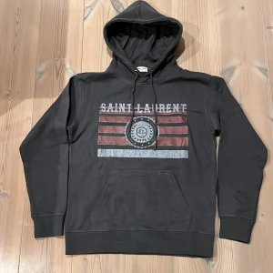 Grå Saint Laurent hoodie - Riktigt fet hoodie från Saint Laurent i storlek M men passar som en S. Snyggt tryck med logga och ränder i rött och silver på bröstet. Vid fler frågor hör gärna av er!🏌️ fraktas inom 24h✅
