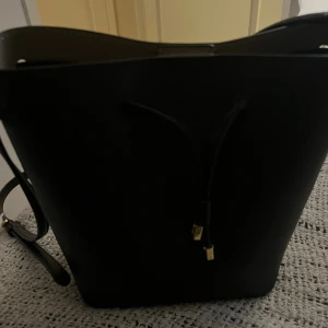 Svart axelväska från Ralph Lauren - Snygg svart axelväska från Ralph Lauren med RL-logga i perforerat mönster. Väskan har justerbar axelrem, snörning upptill och guldfärgade detaljer. Insidan är ljusbeige med praktiska fack och magnetknäppning. Perfekt storlek för allt du behöver ha med dig.