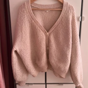 Beige stickad kofta från Kilky Paris - Mysig beige stickad kofta från Kilky Paris med v-ringad hals och två stora knappar framtill. Koftan har en loose passform och långa ärmar, perfekt för lager på lager. Ribbad struktur och mjukt material gör den extra skön att bära.