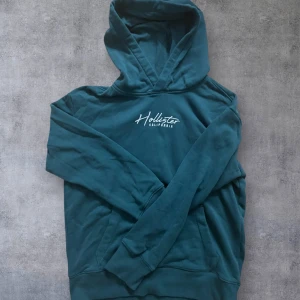 Mörkgrön hoodie från Hollister - Snygg mörkgrön hoodie från Hollister Must-Have Collection med vit broderad logga på bröstet och huvan. Tröjan har klassisk känguruficka och långa ärmar, perfekt för chill dagar. Mjuk bomullskänsla och relaxed passform. Kan passa M.