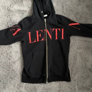 Svart hoodie från Valentino med röd print - Svart hoodie från Valentino med stor röd logoprint på bröstet och armarna. Tröjan har huva, dragkedja framtill och två fickor. Tillverkad i Italien i mjuk bomull. Perfekt för dig som vill ha en statement piece med lyxig vibe.