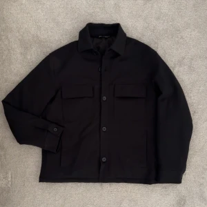 Svart overshirt med fickor - Stilren svart overshirt från Zara med klassisk krage och stora bröstfickor med lock. Jackan har knäppning framtill med stora knappar och raka ärmar. Perfekt för lager-på-lager och en clean look. Storlek M sitter som L 