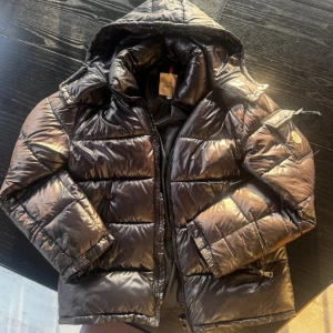 Moncler Jacka  M - Snygg svart pufferjacka från Moncler med glansig finish och huva. Jackan har dragkedja framtill, fickor med dragkedja och en extra ficka med Moncler-logga på ärmen. Perfekt för kalla dagar och riktigt stilren look. Jackan köp på plick vintern 2024. Alla NFC scans fungerar. Är 185 cm och jackan passar perfekt. 