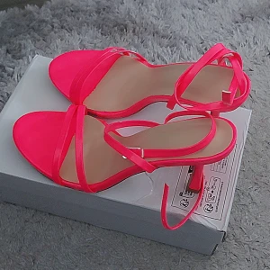 Neonrosa sandaler med klack och remmar - Säljer ett par trendiga neonrosa sandaler med smala remmar och öppen tå. Skorna har en låg klack och justerbara ankelremmar som knyts runt vristen. Perfekta för att sticka ut och ge färg till din outfit. Materialet är syntet med glansig finish. Storlek är asiska 41, i europeiska storlek 40