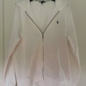 Vit hoodie från Polo Ralph Lauren  - Vit hoodie från Polo Ralph Lauren i storlek XS, det är en barnmodell så därför står det XL. Klassisk design med dragkedja framtill, två fickor och broderad marinblå logga på bröstet. Mjuk bomullskänsla och huva för extra chill vibe. Perfekt för lager-på-lager under höst och vår.
