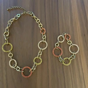 Färgglatt halsband och armband med ringar - Säljer ett set med halsband och armband i guldfärgad metall. Båda har stora cirkelringar dekorerade med glittrande stenar i orange, gul och vit. Kedjorna är chunky och ger en cool statement-look. Perfekt för dig som gillar färg och bling!