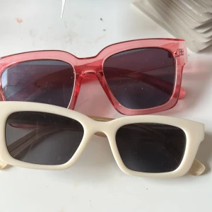 Två trendiga solglasögon i retrostil - Säljer två par solglasögon med rektangulära bågar, ett par i transparent rosa och ett par i matt vit. Båda har mörka glas och cool, chunky design som ger en riktigt modern vibe. Perfekta accessoarer för att lyfta din stil i sommar.