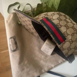 Gucci keps med GG-mönster och röd/grön detalj - Säljer en beige keps från Gucci med klassiskt GG-monogram över hela kepsen och en ikonisk röd/grön rand på sidan. Justerbar rem baktill och svart insida. Kepsen är tillverkad i canvas med detaljer i läder och har en lyxig känsla., Äkta!