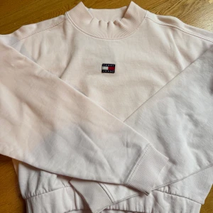 Ljusrosa tröja från Tommy Jeans - Supersnygg ljusrosa sweatshirt från Tommy Jeans med broderad logga på bröstet. Tröjan är i mjukt material med fin passform. Av modellen ”mockneck”. Perfekt för en chill och stilren look. Oanvänd med prislapp kvar. Liknande tröjor ligger på ca 1000kr🩷