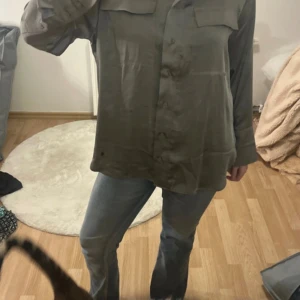 Grå oversized skjorta i silkeslook - Snygg grå skjorta med silkeslen känsla och oversized passform. Skjortan har långa ärmar, två bröstfickor och knäppning framtill. Perfekt för en chill och avslappnad stil.