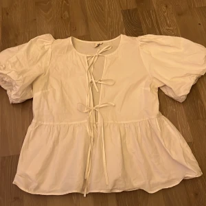 Vit blus med knyt från Nelly - Supersöt vit blus från Nelly med puffiga korta ärmar och flera knytningar framtill. Blusen har en lätt volang nertill och är tillverkad i ett tunt bomullstyg. Perfekt för en romantisk och trendig look. Passar mindre storlekar också 