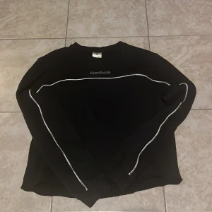 Svart collegetröja tröja från Reebok - Svart collegetröja tröja från Reebok i storlek XS. Tröjan har vita detaljer längs ärmarna och över bröstet samt Reebok-logga framtill. Jag har klippt av tröjan en gång i tiden. 
