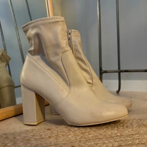Beiga stövletter med klack - Snygga beiga stövletter med hög blockklack och rund tå. Skorna har dragkedja på insidan och är gjorda i ett slätt syntetmaterial som ger en clean look. Perfekta för att lyfta vilken outfit som helst.