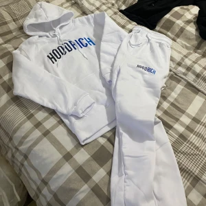 Vit hoodie från Hoodrich med blå text - Säljer en fräsch vit hoodie från Hoodrich med stor HOODRICH-logga i svart och blått på bröstet. Hoodien har huva med snörning, magficka och ribbade muddar. Perfekt för chill eller streetwear. Materialet är mjuk bomull och polyesterblandning. kan passa XS