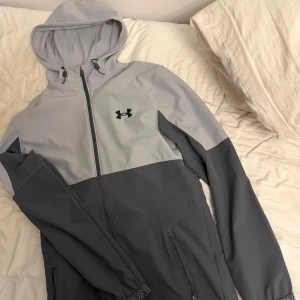 Grå vindjacka från Under Armour - Snygg tvåtonad grå vindjacka från Under Armour med huva och hel dragkedja framtill. Jackan har långärmade ärmar, två sidofickor med dragkedja och logga på bröstet. Perfekt för dig som gillar sportig stil och vill ha något lätt och funktionellt.