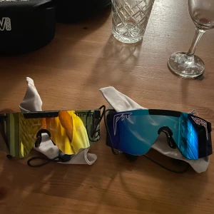 2 par Pit Viper 2000 sportglasögon - Två par äkta Pit Viper 2000 sportglasögon med spegellinser, ett par i gult och ett i blått. Båda har breda, rektangulära linser och svart båge. Perfekta för dig som vill sticka ut och få en futuristisk look. Kommer med originalfodral.