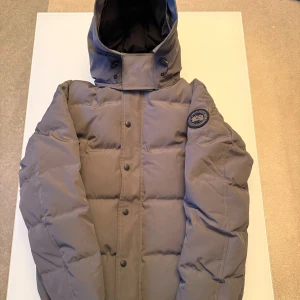 Grå dunjacka från Canada Goose - Säljer en grå dunjacka från Canada Goose med huva och snyggt märke på ärmen. Jackan har stora fickor med dragkedja och knappar framtill. Perfekt för kalla dagar och har en clean, puffad look som är riktigt nice.    Adda min snap för mer bilder !!!💥💥💥