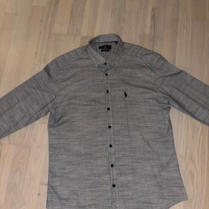 Grå skjorta från Polo Ralph Lauren - Snygg grå skjorta från Polo Ralph Lauren med diskret rutig struktur och klassisk krage. Skjortan har svarta knappar hela vägen och lång ärm med manschett. Ikoniska Polo-loggan broderad på bröstet. Perfekt för dig som gillar stilrena detaljer.