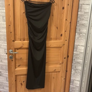Svart supersnygg långklänning - Säljer en svart, axelbandslös långklänning med tight passform. Klänningen har snygg rynkdetalj på ena sidan och är gjord i ett stretchigt, mjukt material som sitter figurnära. Perfekt för dig som vill ha en stilren och elegant look.