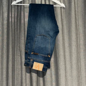 Blå Nudie Jeans - Säljer ett par klassiska blå jeans från Nudie Jeans med snygga kontrastsömmar och bakficka med broderad detalj. Jeansen har en rak passform och är tillverkade i slitstarkt bomullsdenim. Perfekta för dig som gillar stilrena och tidlösa jeans.