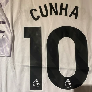Manchester United Cunha #10 fotbollströja - Säljer en Manchester United fotbollströja, Adidas i slim fit, med Cunha och nummer 10 på ryggen. Tröjan är vit med ljuslila grafiskt mönster, har korta ärmar och V-ringad krage med lila kant. Materialet är lätt och andas, perfekt för match eller träning.
