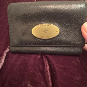 Svart plånbok från Mulberry i skinn - Snygg svart plånbok från Mulberry i äkta skinn med klassisk guldfärgad metallplakett framtill. Plånboken har flera fack för kort och två fack med dragkedja för mynt. Perfekt storlek för att ha med sig överallt och stilren design.