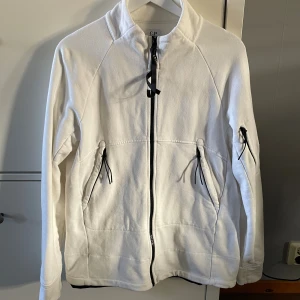 Vit C.P. Company tröja stl. M - Vit Tröja från C.P. Company med svarta detaljer och dragkedjor. Jackan har hög krage, två fickor framtill och en ärmficka med den klassiska C.P. Company-linsen