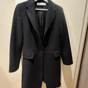 Svart kappa från Zara med ull - Stilren svart kappa från Zara med klassisk krage och snygga fickdetaljer. Tillverkad i ull från Manteco, med knappar framtill och på ärmslutet. Lite nopprig men går lätt att få bort! I bra kvalité. Nypris 1799. Jag upplever storleken som liten