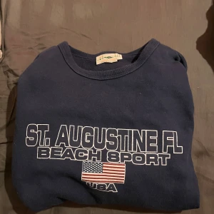 Marinblå sweatshirt St. Augustine FL - Säljer en marinblå sweatshirt från Retro Sport med tryck 'St. Augustine FL Beach Sport' och amerikansk flagga på bröstet. Klassisk rund halsringning och avslappnad passform. Perfekt för dig som gillar sportig och casual stil.