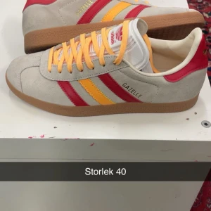 Adidas Gazelle sneakers grå/röd/gul - Säljer ett par Adidas Gazelle sneakers i grå mocka med röda och gula ränder på sidorna. Skorna har klassisk gummisula, rund tå och snörning med gula skosnören. Perfekta för dig som gillar retrostil och färgkombinationer.