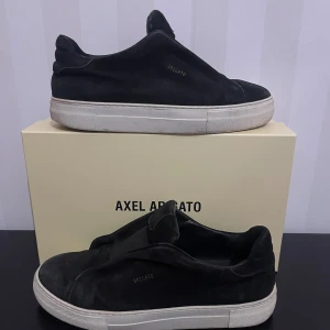 Svarta Axel Arigato i mocka - Svarta sneakers från Axel Arigato i mjuk mocka med vit platt sula. Minimalistisk design utan snörning, med elastisk panel över vristen och diskret logga på sidan. Insidan är fodrad med skinn för extra komfort. Perfekt för dig som gillar stilrena och cleana skor. Skorna är rätt så använda så lite smutsiga men det går alltid att lösa annars inga andra skador. 