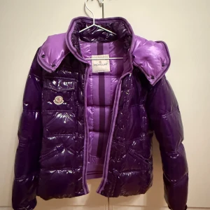 Lila pufferjacka från Moncler - Säljer en glansig lila pufferjacka från Moncler med avtagbar huva. Bra skick förutom att etiketten är påväg att lossna på insidan. Köpt för flera år sedan så en gammal modell. Står inte storlek men skulle gissa på S.