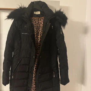 Svart lång pufferjacka Roco Baroco - Svart pufferjacka från Roco Baroco med fluffig fuskpäls på huvan och leopardmönstrat foder. Jackan har flera dragkedjor, långa ärmar och en snygg, quiltad design. Perfekt för kalla dagar när du vill vara både varm och stilren.
