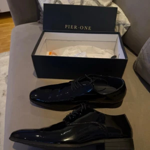 Svarta lackskor från Pier One - Säljer ett par svarta, glansiga lackskor från Pier One. Skorna har spetsig tå, snörning och låg klack. Perfekta för dig som vill ha en stilren och klassisk look. Materialet är syntet med en riktigt snygg finish. Bara använda 1 gång men inget man märker av.