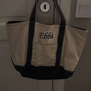  Tote Stories - Stor och rymlig shoppingväska i beige, bomull med breda svarta handtag och detaljer. Framsidan har ett broderat namn 'Ebba' och en praktisk ytterficka. Perfekt för att bära allt du behöver till skolan eller stan.