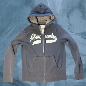 Mörkblåhoodie Abercrombie & Fitch - Snygg mörkblå hoodie från Abercrombie & Fitch med stor vit broderad logga på bröstet. Tröjan har huva med grått foder, dragkedja framtill och två fickor. Materialet är mjuk bomull och polyester, perfekt för chill dagar. Klassisk streetstil med ribbade muddar. Enda defekten är att den saknar snören. 