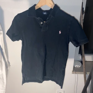 Mörkblå pikétröja från Polo Ralph Lauren - Klassisk mörkblå pikétröja från Polo Ralph Lauren med korta ärmar och krage. Tröjan har en rosa broderad logga på bröstet och är tillverkad i mjuk bomull. Perfekt för en stilren och avslappnad look. 149 kr