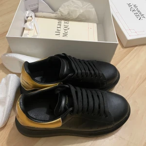 Svarta sneakers Alexander McQueen - Snygga svarta sneakers från Alexander McQueen med metallicguld detalj på hälen. Skorna har platt sula, rund tå och klassisk snörning. Tillverkade i skinn med en chunky siluett som ger en cool och modern vibe. Perfekta för dig som gillar statement-skor.