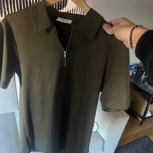  pikétröja från NN07 - Snygg olivgrön pikétröja från NN07 med kort ärm och klassisk krage. Tröjan har en halv dragkedja framtill som ger en modern touch. Tillverkad i mjuk bomull som känns skön mot huden. Perfekt för dig som gillar stilrena och enkla plagg. Aldrig använd då den var lite stor för mig. S men skulle säga att den är mer som en M